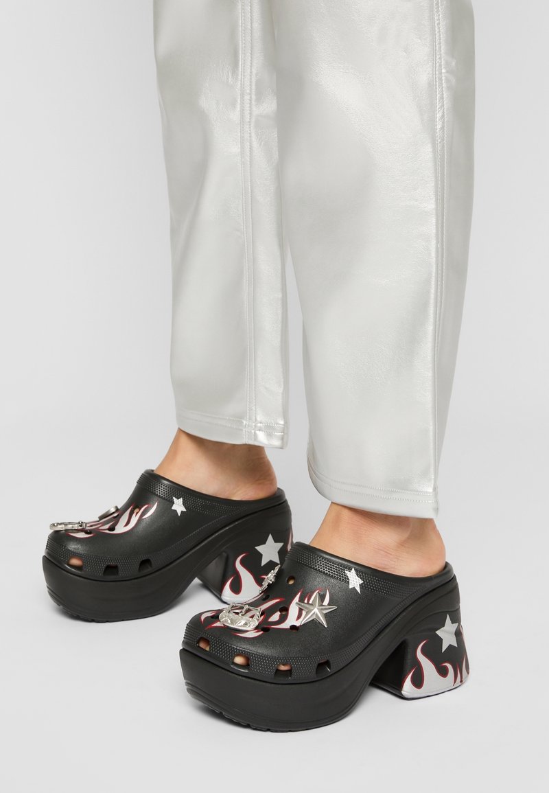 Sabots à plateforme noirs avec des motifs de flammes et d'étoiles portés avec un pantalon blanc, montrant les chevilles et les pieds sur un fond clair.
