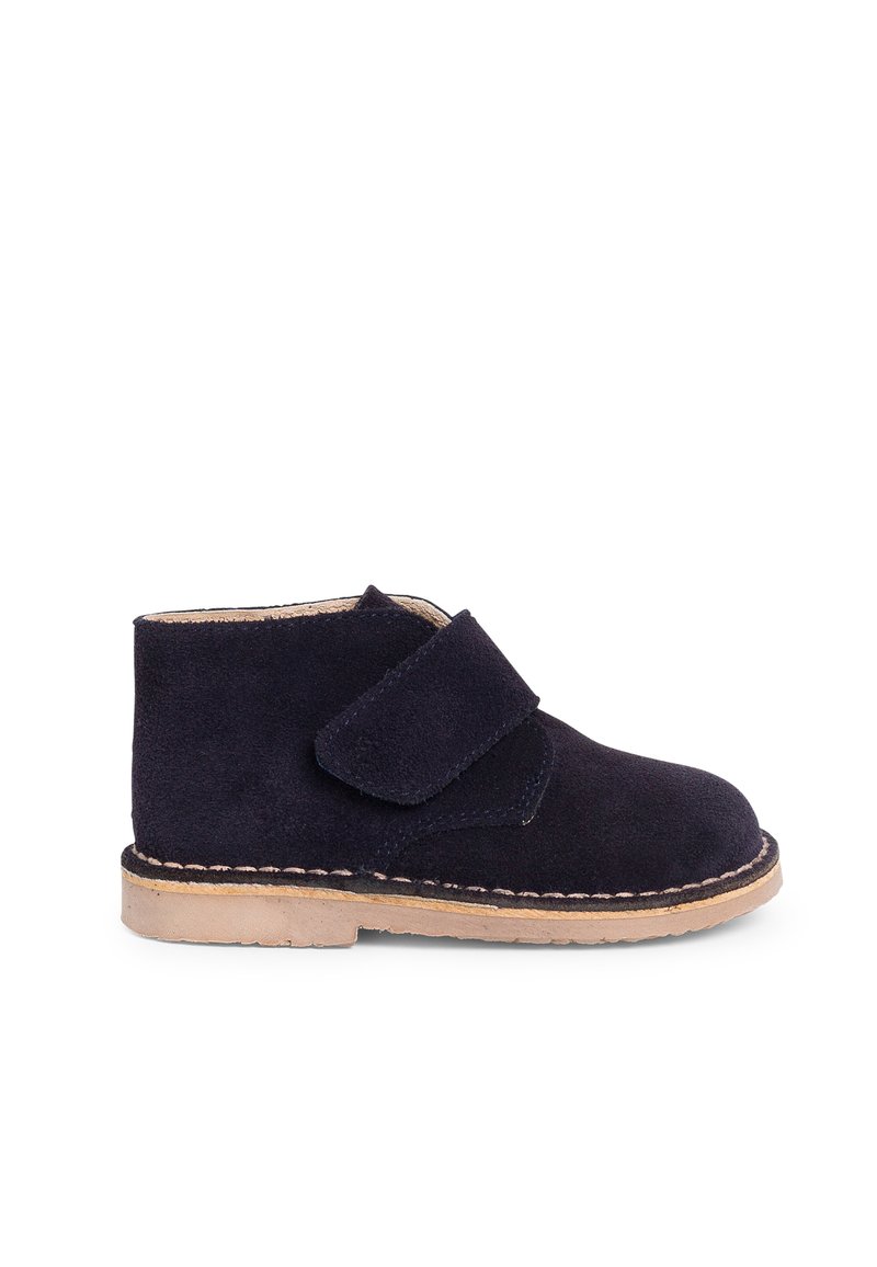 Bottines en daim bleu marine avec une sangle Velcro, bout arrondi et semelle brun clair avec un détail cousu. Texture douce et design simple.