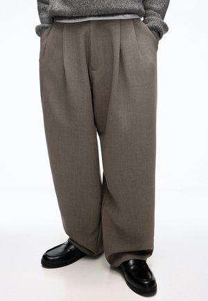 Persoon die losse bruine broek, zwarte schoenen en een grijze trui draagt, met handen in de zakken tegen een witte achtergrond.