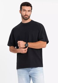 Černé bavlněné tričko s uvolněným střihem, kulatým výstřihem a krátkými rukávy, spárované s světlomodrými džíny a černými hodinkami na zápěstí.