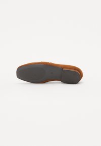 Chaussure en daim marron avec un bout arrondi et un talon en caoutchouc noir bas. La semelle présente un motif de grip texturé.