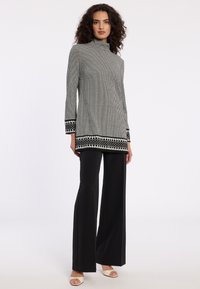 Zwart-wit houndstooth tuniek met een hoge halslijn, lange mouwen en geweven patroonrand. Gecombineerd met zwarte brede broekspijpen en sandalen.