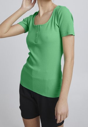 Frau trägt ein eng anliegendes, kurzärmeliges, grünes, geripptes Shirt mit Knöpfen und schwarze Shorts, posiert mit einem Arm erhoben in der Nähe ihres Gesichts.