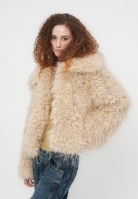 Μπεζ τζάκετ από συνθετική γούνα με φαρδιά γιακά και αφράτη υφή, με μακριά μανίκια και κοντό σχέδιο, συνδυασμένο με κίτρινο τοπ και τζιν.