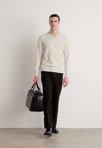 Beige V-halsgenser, svarte bukser og svarte sko. Holder en svart skinnveske med håndtak. Minimalistisk design, glatt tekstur.