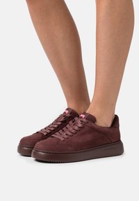 Wildleder-Sneaker in Burgunderrot mit runder Spitze, flacher Sohle und Schnürdesign. Mit rotem Logo an der Ferse und glatter Textur.