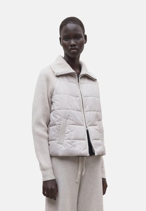 Mujer de piel oscura con chaleco acolchado beige claro con cremallera, mangas y cuello de punto acanalado, combinado con pantalones beige claro a juego con cordón.