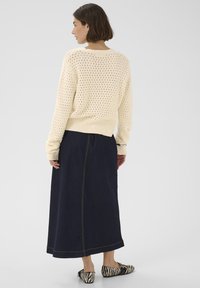 Cremefarbener Strickpullover mit offenem Maschenmuster, kombiniert mit einem langen marineblauen Jeansrock und schwarz-weiß gestreiften flachen Schuhen.