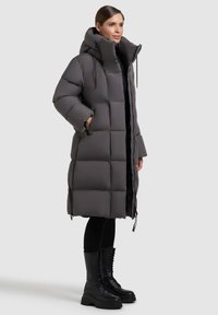 Abrigo acolchado gris con cuello alto, diseño acolchado y detalles ajustables. Combinado con botas de combate negras con cordones y leggings negros.