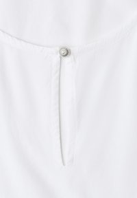 Détail de vêtement blanc avec un bouton décoratif rond en argent au col et une petite ouverture fendue verticale en dessous.