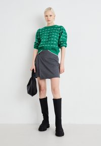 Suéter verde de punto con patrón texturizado, falda envolvente gris, botas negras hasta la rodilla y un bolso negro. Diseño minimalista y moderno.