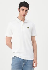 Vit polo-shirt i bomull, med en gul broderad fjärilslogotyp på vänster bröst, klassisk krage och korta ärmar.