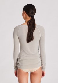 Helles graues Langarmshirt, enganliegend mit rundem Ausschnitt, gerafften Seiten und weichem Material. Getragen mit Unterwäsche in neutralen Farben.