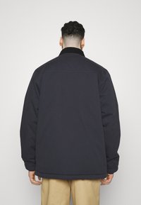 Donkerblauwe jas met een zachte textuur, voorzien van een kraag en een losse pasvorm. Zichtbare stiksels bij de schouders en de onderrand.