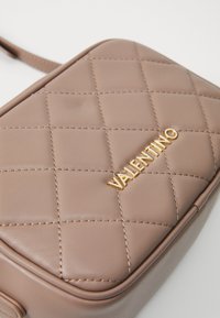 Bežinis siuvinėtas odinis per petį nešiojamas krepšys su auksiniu VALENTINO logotipu ant baltos fono.