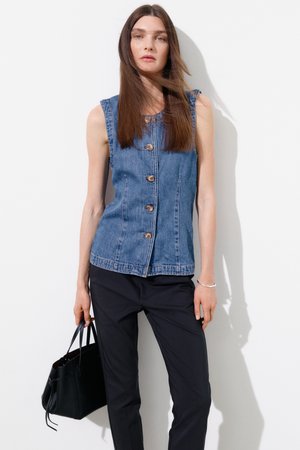 Kobieta z długimi brązowymi włosami, ubrana w bezrękawnik dżinsowy z guzikami, czarne spodnie, trzyma czarną torebkę, stoi na białym tle.