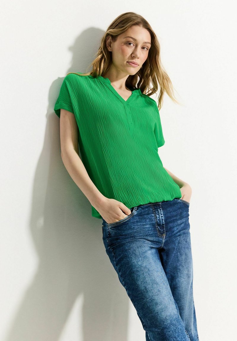 Top verde, plissettato, a maniche corte con scollatura a V, abbinato a jeans in denim blu. Il top ha una texture liscia e i jeans presentano una vestibilità classica.