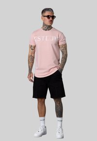 Helles rosa T-Shirt mit "ESTEEM" Grafik, schwarze Shorts, weiße Sneakers und Sonnenbrille. Auffällige Tattoos an Armen und Beinen sichtbar.