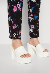 Zuecos de plataforma gruesa blanca con una tira ancha y diseño de punta abierta, llevados con pantalones negros florales que presentan patrones en rosa, blanco y azul.