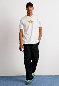 Vit t-shirt med en gul logotyp, kombinerad med svarta, löst sittande byxor och gröna sneakers. Modellen går mot en enfärgad bakgrund.