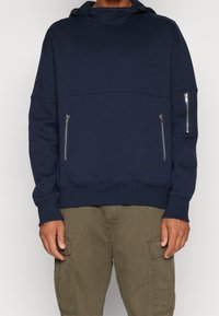 Felpa blu navy con colletto alto, dotata di polsini e fondo a costine, due tasche laterali con zip e una vestibilità ampia.