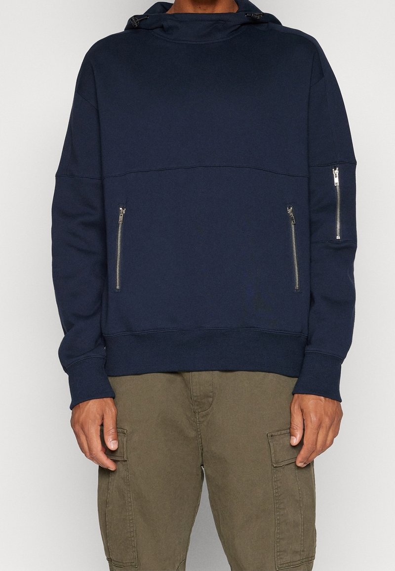 Felpa blu navy con colletto alto, dotata di polsini e fondo a costine, due tasche laterali con zip e una vestibilità ampia.