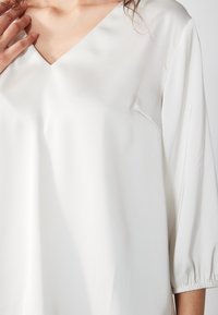 Blouse blanche avec col en V et manches trois-quarts. Tissu lisse et léger avec une subtile brillance et des poignets élastiqués.