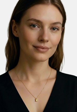 Junge Frau mit heller Haut und braunen Haaren, die ein schwarzes Oberteil und eine goldene Halskette mit einem "Z"-Anhänger trägt, lächelt sanft vor einem schlichten Hintergrund.