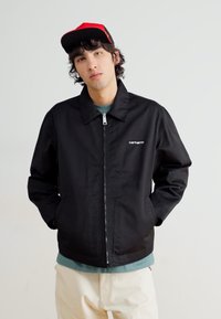 Veste noire avec une fermeture éclair à l'avant et un col pointu. Présente un logo Carhartt sur le côté gauche. Portée sur une chemise bleu clair.