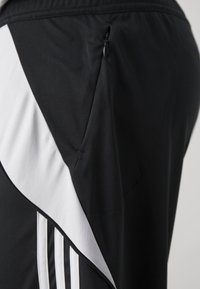 Gros plan d'un pantalon de sport noir avec une bande diagonale blanche et des bandes blanches verticales sur le côté, avec une poche zippée.