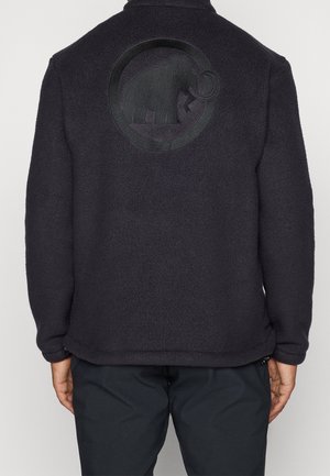 Person trägt eine schwarze Fleecejacke mit eingeprägtem Mammut-Logo auf dem oberen Rücken, kombiniert mit dunkler Hose vor neutralem Hintergrund.