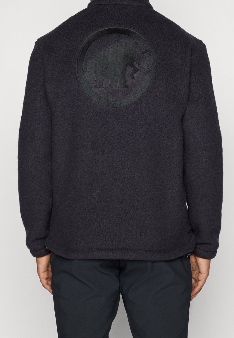 Person trägt eine schwarze Fleecejacke mit eingeprägtem Mammut-Logo auf dem oberen Rücken, kombiniert mit dunkler Hose vor neutralem Hintergrund.