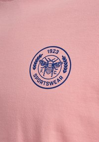 Circulair blauw sportkledinglogo op roze stof met een illustratie van een bij, laurier takken en tekst "1923 F.C. SPORTSWEAR."