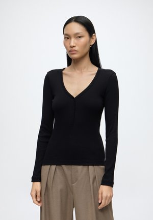 Jeune femme aux longs cheveux noirs porte un haut noir ajusté à col en V et manches longues ainsi qu'un pantalon beige ample devant un fond uni clair.