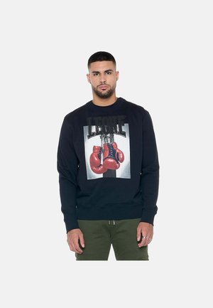 Uomo che indossa una felpa nera con grafica di guantoni da boxe rossi e testo "LEONE 1947", abbinata a pantaloni verde oliva, in piedi su sfondo bianco.