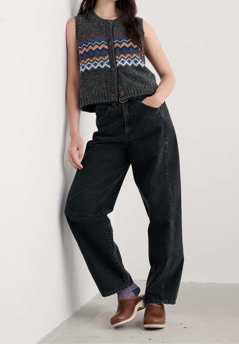 Personne appuyée contre un mur portant un gilet en tricot sans manches à motifs, un jean noir taille haute, des chaussettes violettes et des sabots marron.