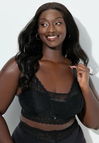 Svart spetsbralett med vågiga kanter, genomskinlig underdel och dekorativ knappdetalj. Modellen har vågigt hår och naturlig makeup som kompletterar utseendet.