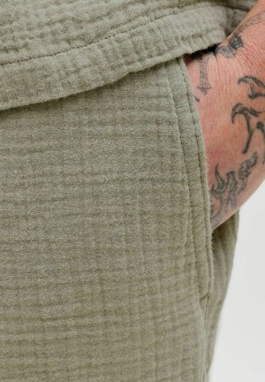 Olive green, textured fabric showing a quilted pattern. Close-up of a pocket detail on a garment with visible seam and soft texture.