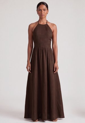Kobieta stojąca, ubrana w długą, bezrękawnikową, ciemnobrązową sukienkę maxi z dopasowanym gorsetem i zwiewną spódnicą na jednolitym tle.