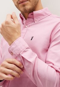 Camicia button-up rosa chiaro con colletto, dotata di dettagli con bottoni e un piccolo emblema di aragosta scura sul petto.