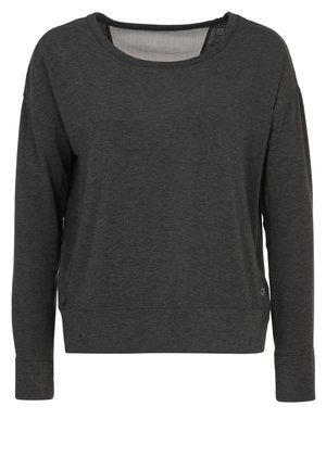 Persona con camiseta de manga larga gris oscuro, leggings cortos negros y zapatillas deportivas negras y moradas, de pie sobre un fondo claro.