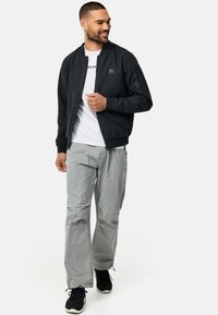 Svart bomberjakke med glidelås foran og lommer, kombinert med lysegrå bukser og svarte sneakers. Modell som står på en enkel bakgrunn.