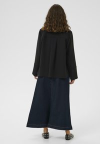 Juodas ilgas rankovėmis marškinis su klostėta nugara, derinamas su ilgu džinsiniu sijonu, turinčiu apvadu ir kontrastingą siuvinėjimą, nešiojamas su smeigėmis puoštomis bateliais.