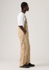 Άνδρας με πλεγμένα μαλλιά, φορώντας μπεζ φορμά συνολικά, λευκό κοντομάνικο πουκάμισο, μαύρα παπούτσια, στέκεται πλευρικά με τα χέρια στις τσέπες μπροστά σε απλό φόντο.