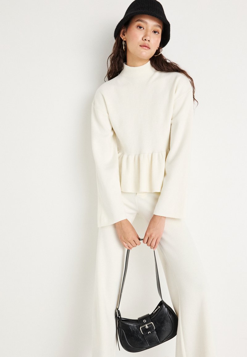 Top bianco a maniche lunghe con collo alto e maniche svasate, dotato di un orlo peplum. Abbinato a pantaloni bianchi e una borsa nera con dettaglio a fibbia.