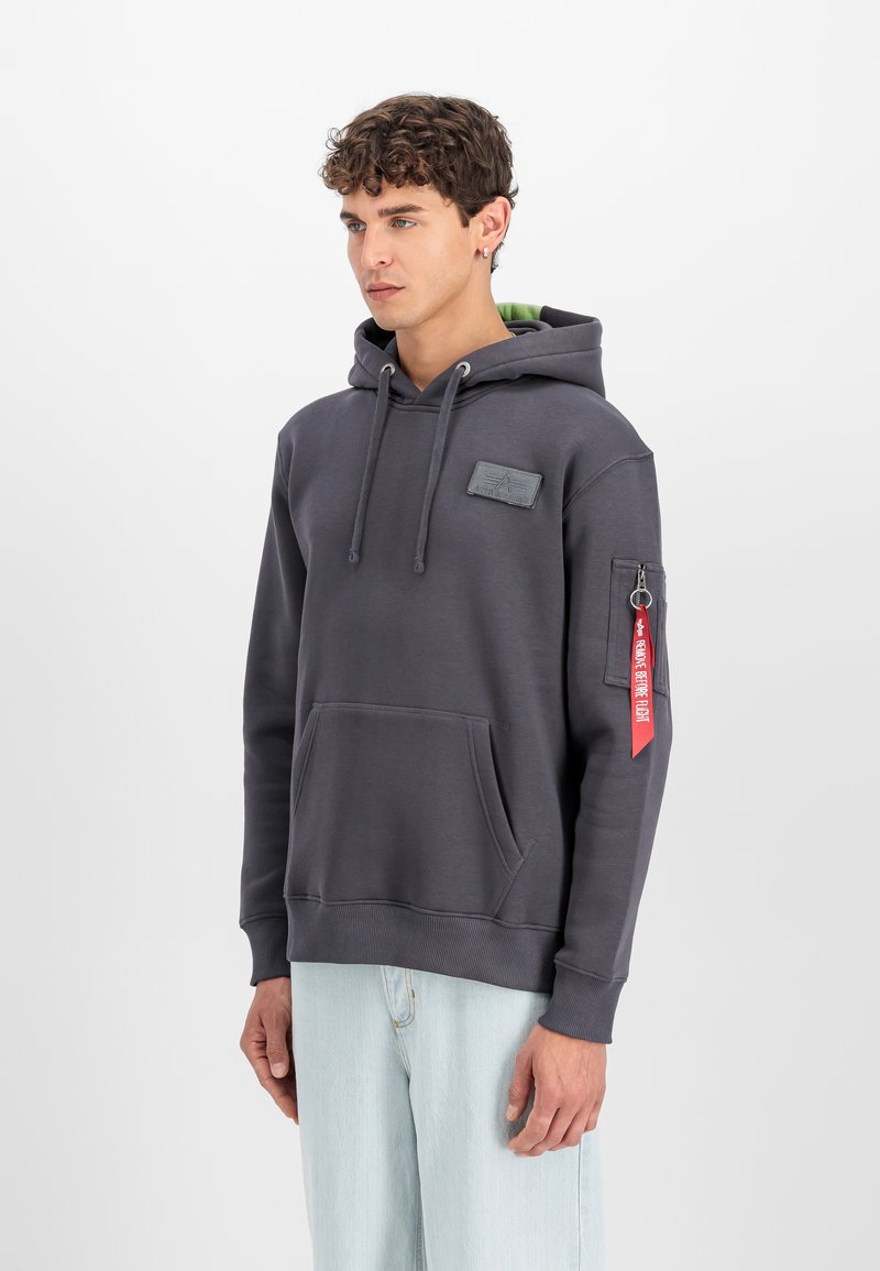 Jonge man met krullend haar, die een donkergrijze capuchonsweater draagt met een voorzak en een rood label op de linkermouw, staand tegen een witte achtergrond.