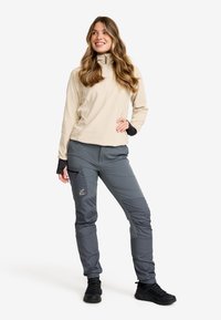 Leende kvinna med långt vågigt hår, klädd i en beige zip-up fleece, grå cargobyxor och svarta sneakers, stående med ena handen på höften.