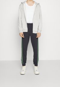 Grå zip-up huvtröja, vitt linne, marinblå mjukisbyxor med gröna sidor och vit logotyp, kombinerat med vita högklackade sneakers.