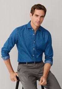 Džínová košela na gombíky v tmavomodrej farbe, s klasickým golierom a vyhrnutými rukávmi, v kombinácii so sivými nohavicami a tmavým opaskom.