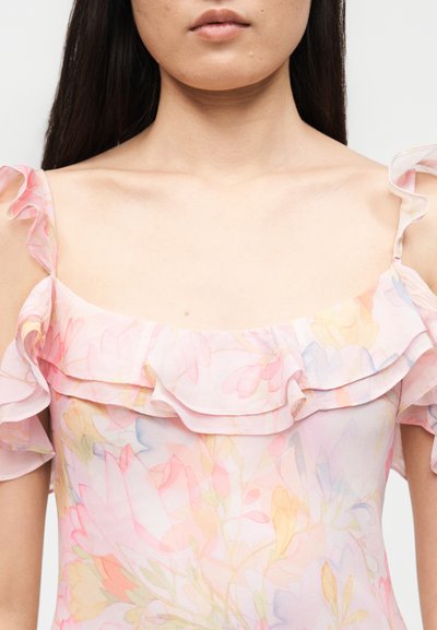 Γυναίκα που φοράει παστέλ ανθισμένο φόρεμα με σούρες στα μανίκια που αφήνουν τους ώμους ακάλυπτους και τετράγωνο ντεκολτέ, εμφανίζοντας τον λαιμό και το ανώτερο τμήμα του στήθους.